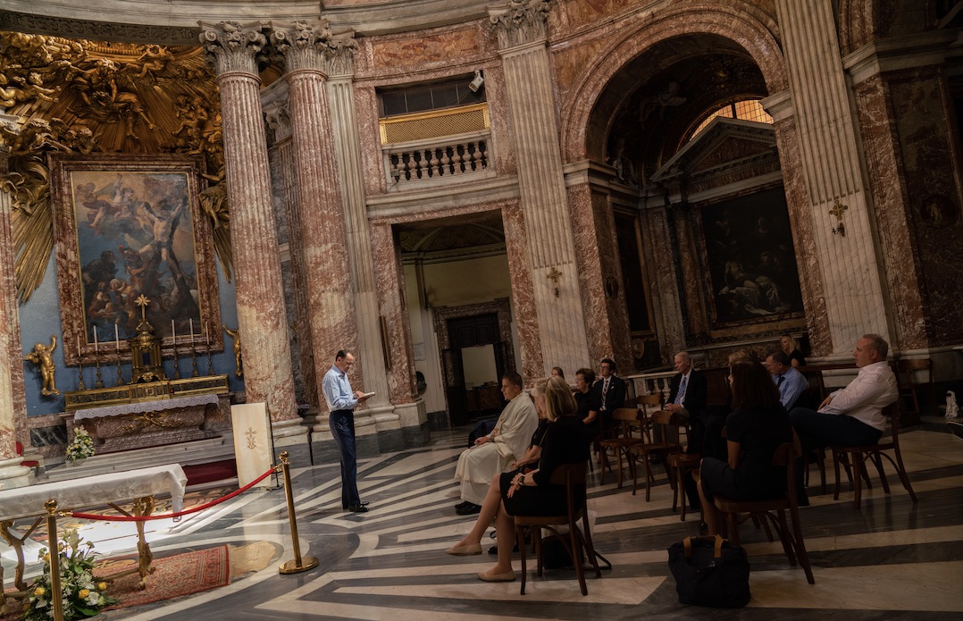 A man reads from a book to a seated audience in a vast church.