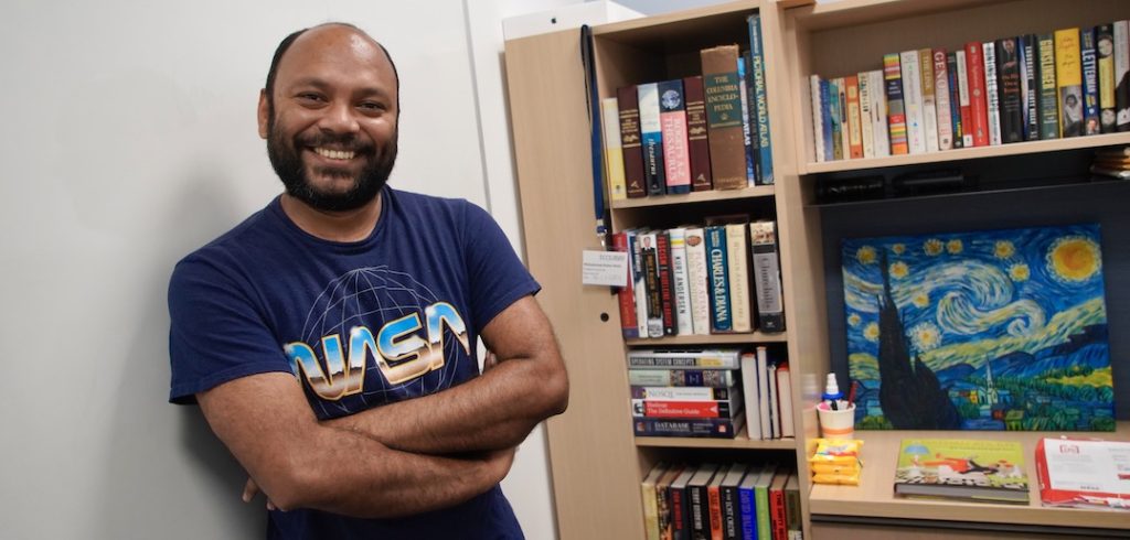 A man wearing a blue NASA shirt crosses his arms in front of his chest and smiles in front of a bookshelf and painting.