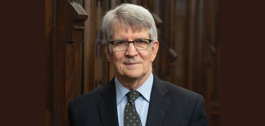 A gray-haired man wearing a suit smiles in front of a wooden panel.