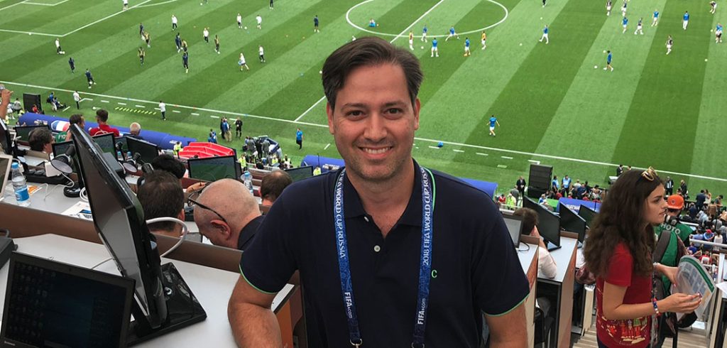 Journalist and Fordham graduate Clemente Lisi in the press area at a match during the 2018 World Cup in Russia, with a match being played on the field behind him