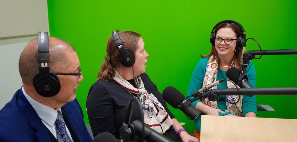 Three seated people wearing headphones speak into microphones.