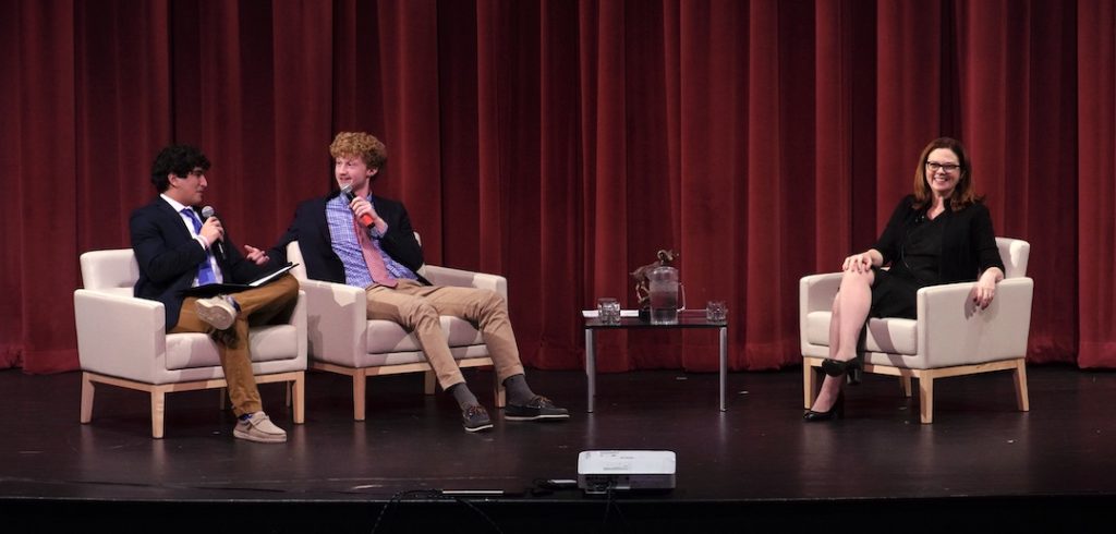 Three seated people in white armchairs sit and laugh in front of a long red curtain on a stage.