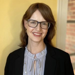 A woman with glasses smiles in front of a yellow wall.