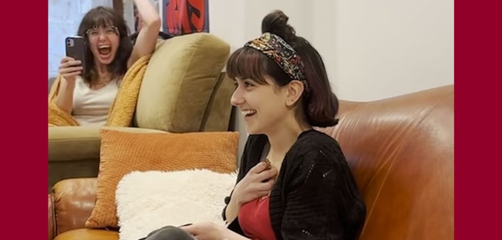 A young woman holds a hand to her chest in surprise while sitting on a couch, while a young woman in the background cheers in support.