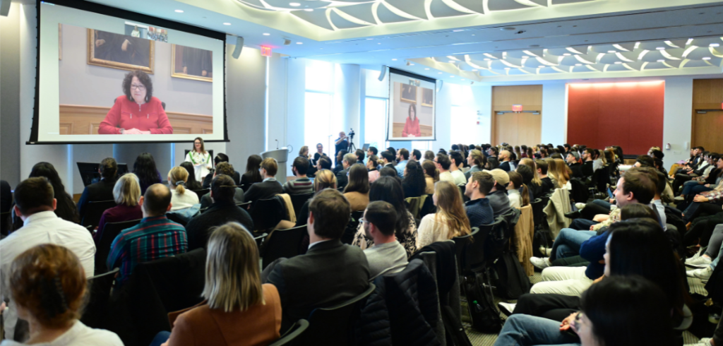 Conference room with a group of people sitting in chairs looking at a screen with a woman talking.