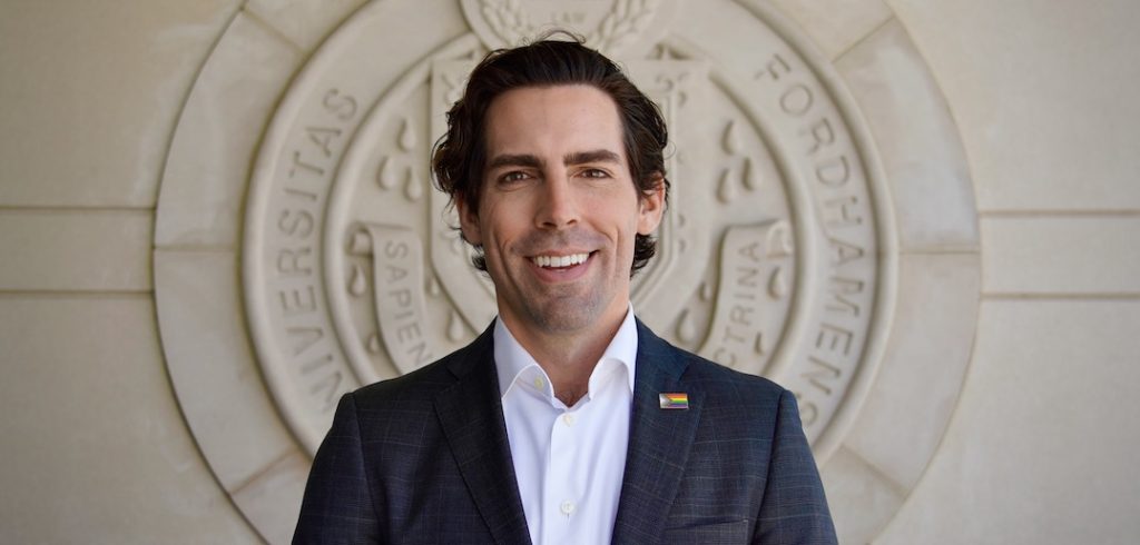 A man wearing a blazer and a rainbow pin smiles.