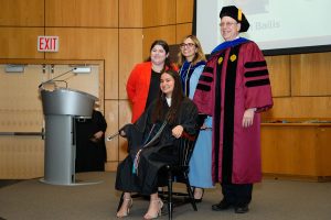 A female student seated in a chair on stage, surrounded by three administrators