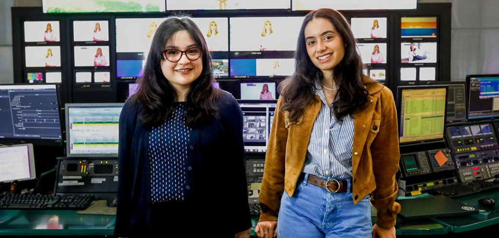 Two women leaning against a video control board, with numerous monitors behind them them