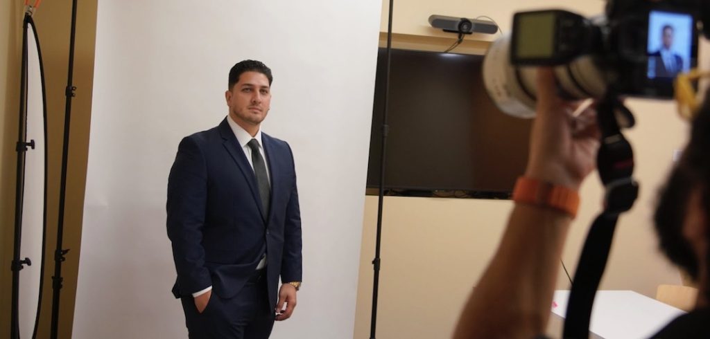 A student veteran wearing a suit smiles for a camera while standing in front of a professional backdrop.