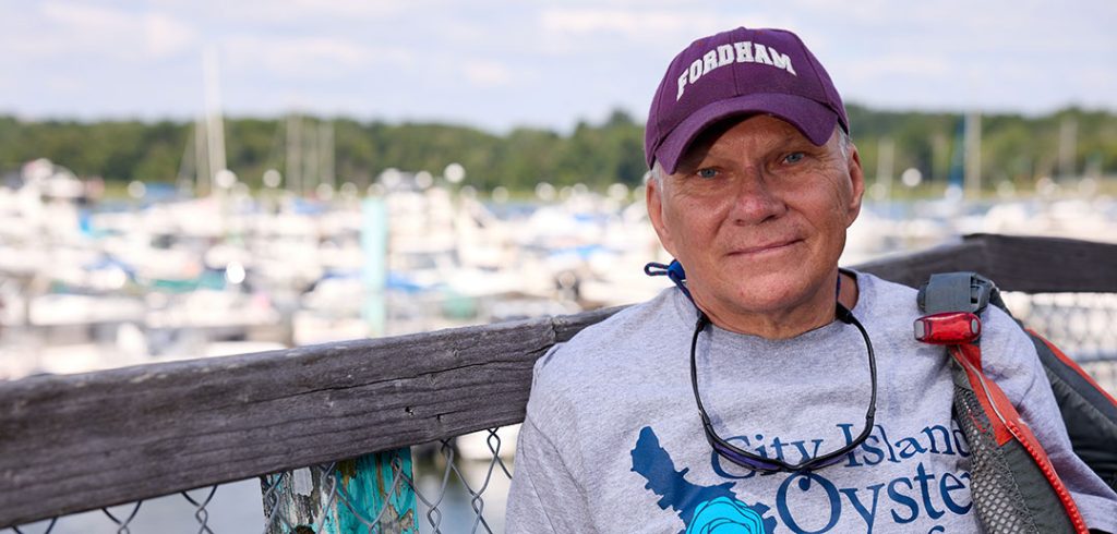 A man wearing a Fordham hat and a City Island Oyster Reef T-shirt on a dock with boats on the water in the background