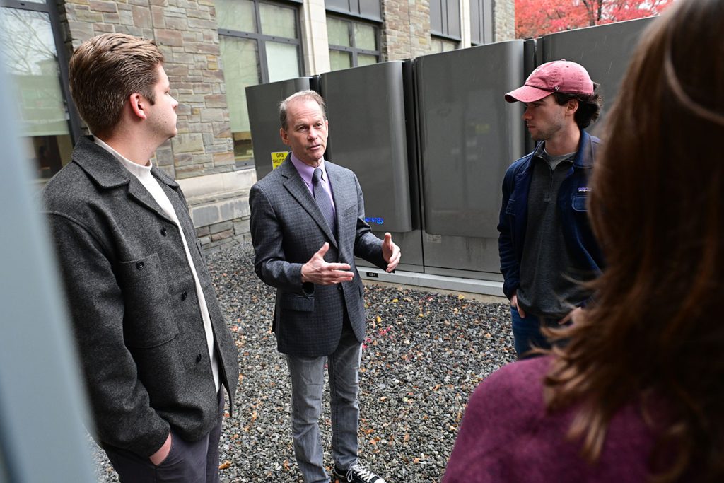 Students stand in a circle, listening to a man standing next to a man with his hands out in front of him.