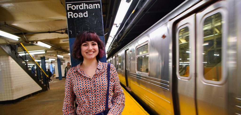 A woman stands against a pillar on a Fordham Road subway platform, a car coming into the station next to her