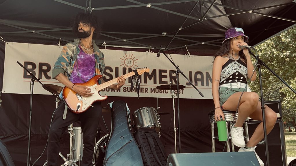 Male presenting individual with electric guitar and female presenting individual on microphone. On stage at the Bronx Summer Jam.
