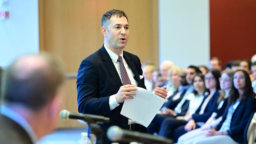 A man wearing a suit and holding a piece of paper speaks to a crowd seated in front of him.