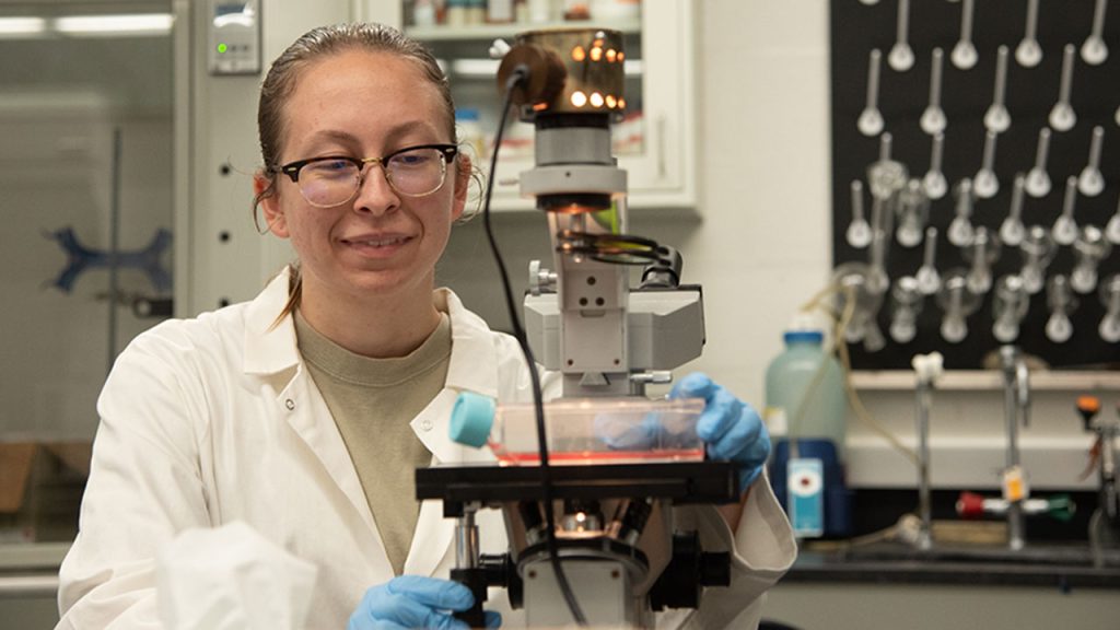 Mary Biggs in science lab looking down at microscope. She is in a white lab coat.