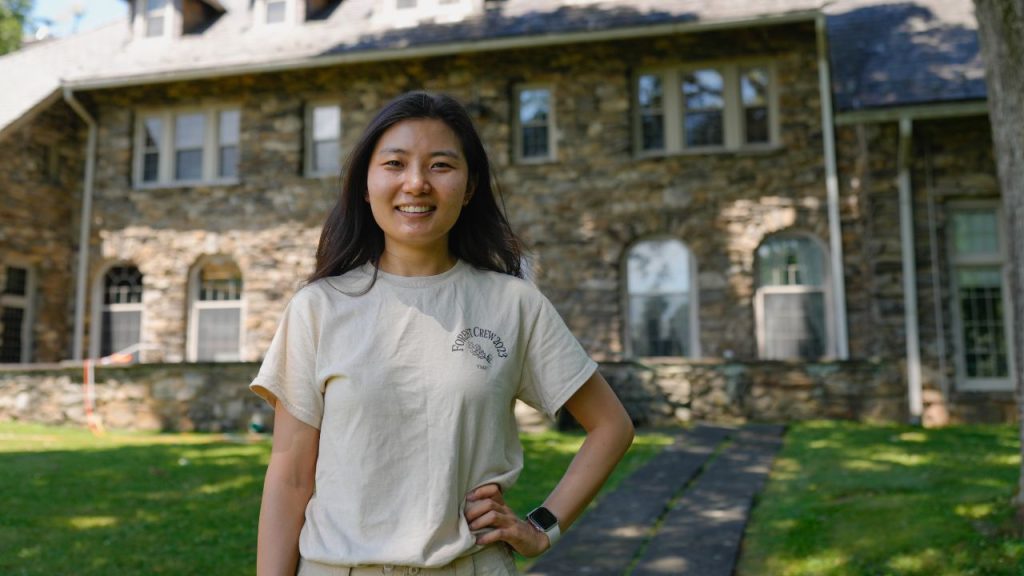 A woman stands in front of a stone building with one hand on her hip.