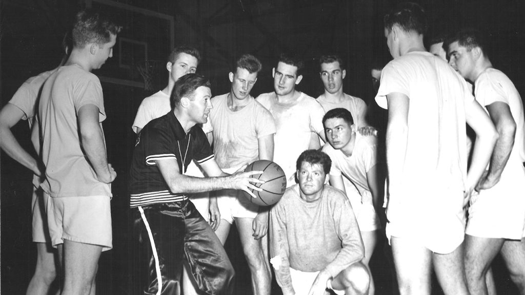 Basketball players gather around and look at a coach holding a basketball in the middle of the huddle