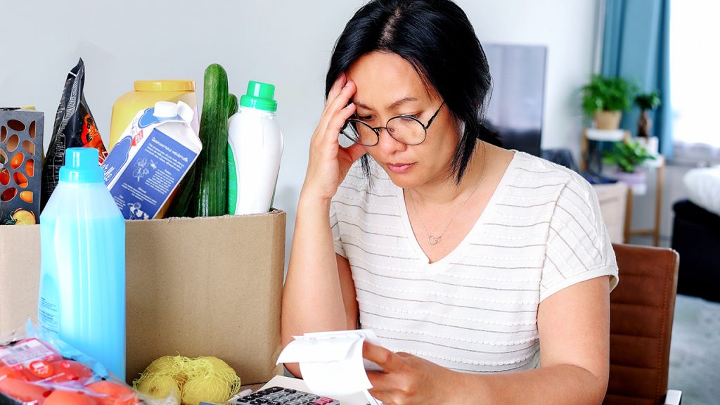 A woman seated at a table with groceries, looking anxiously at her receipt