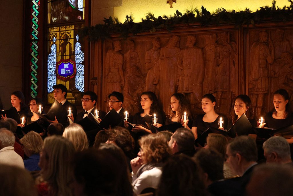 Ten members of the chorus sing while standing alongside the walls of the University Church, while holding candles.