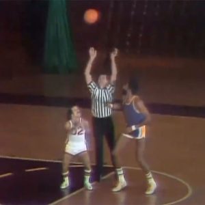 A referee throws a basketball up for a jump ball between two players, one significantly taller than the other