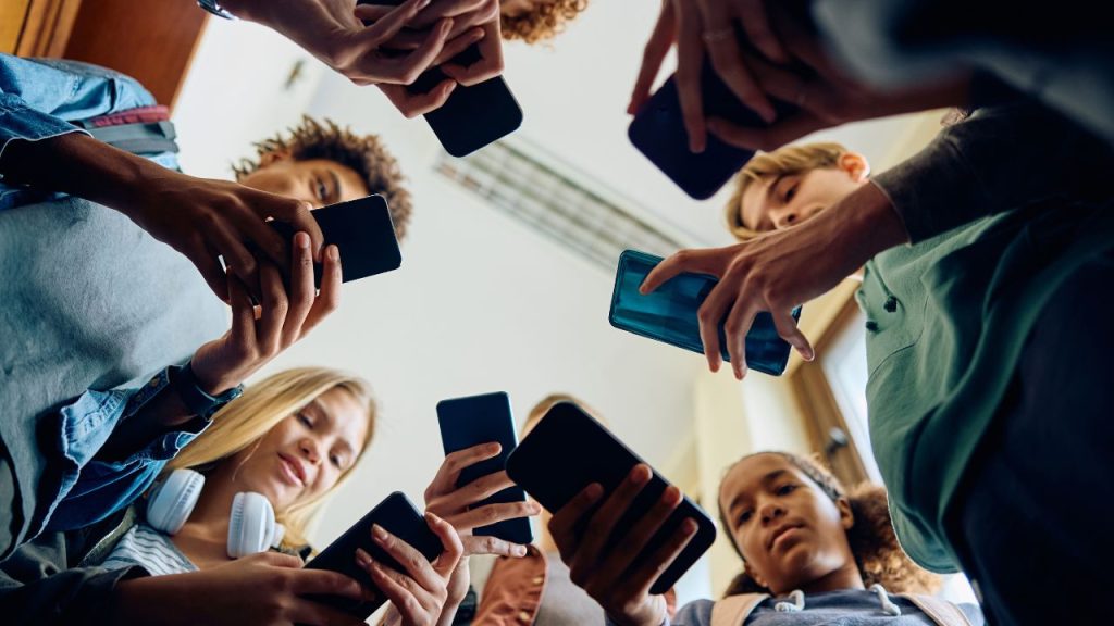 A group of teens stand in a circle looking down at their cell phones, representing the New York cell phone ban in schools proposed by Governor Kathy Hochul's executive budget for fiscal year 2026.