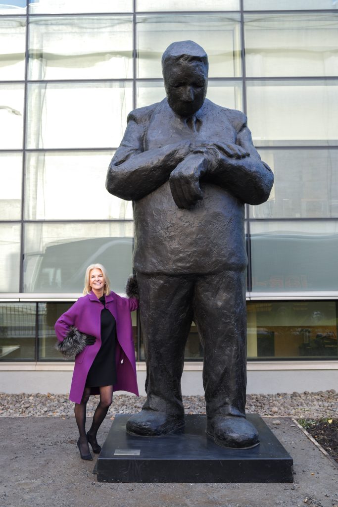 Fordham Trustee Kim Bepler stands in a purple coat leaning against a monumental sculpture of a male figure by Jim Rennert