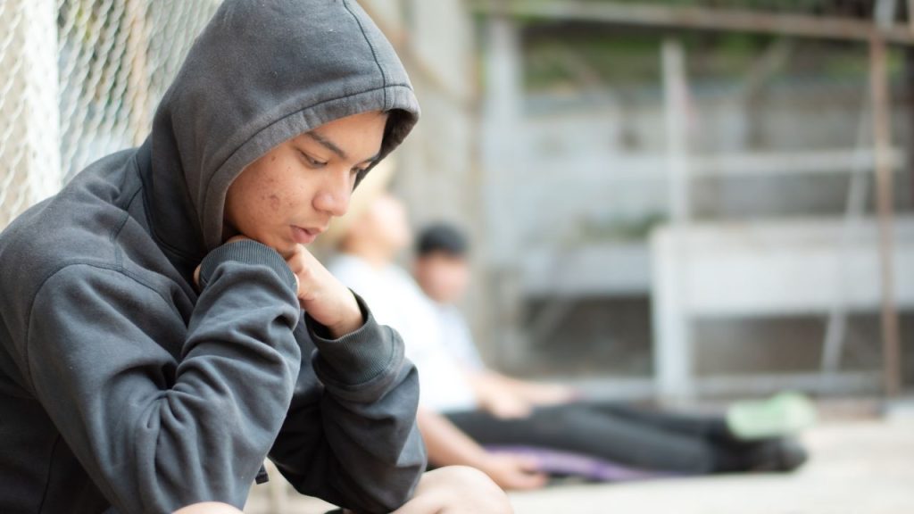 an adolescent sits with his back to a chainlink fence, representing traumatized kids in the juvenile justice system