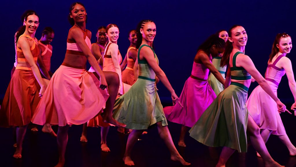 Women dancers fdressed in multi-colored dresses face the audience with their hands pointed downward.