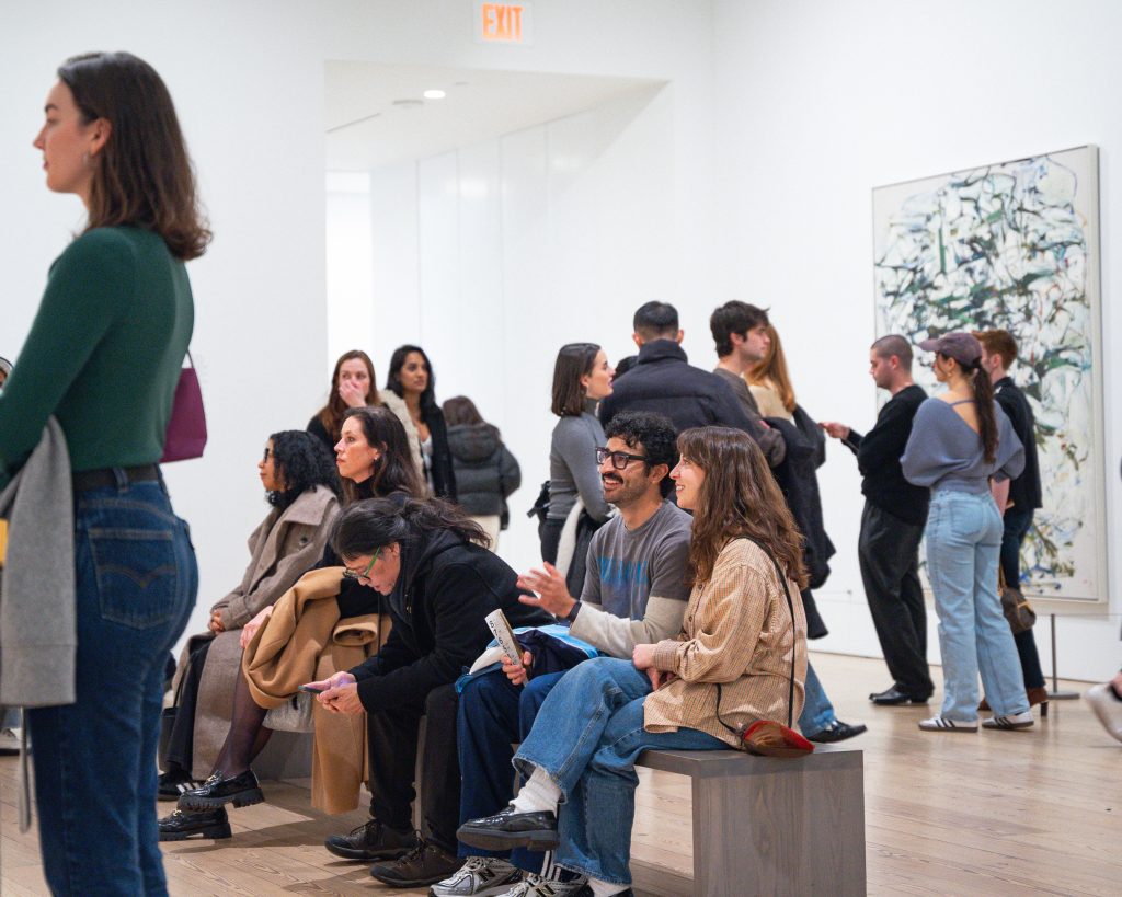 Group of people in Whitney Museum, some looking at art on wall and some sitting down.
